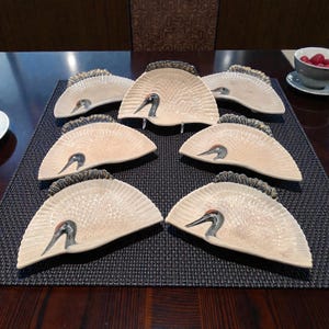 May include: A set of seven semi-circular ceramic dishes, each featuring a sculpted crane head with a red crest and a textured fan-shaped body in a cream color. The edges of the fan are detailed with dark, feathered patterns in a relief design.