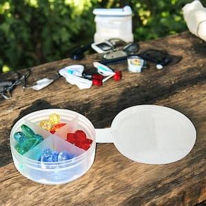 May include: A clear plastic pill box with a lid. The pill box is divided into four sections. Each section contains colorful plastic beads.