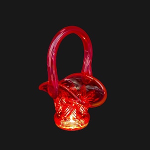 May include: A vibrant red glass basket with a curved handle. The basket has a textured base and a rounded body. The handle arches gracefully, and the glass has a glossy finish. The basket is set against a black background.