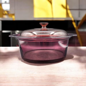 May include: A clear glass casserole dish with a purple tint, featuring a matching lid and a small handle. The dish sits on a wooden surface, showcasing its elegant design. The casserole dish is perfect for baking and serving.