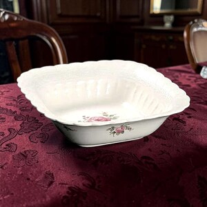 May include: A square white ceramic serving bowl with scalloped edges and a textured rim. The interior and exterior feature a delicate floral pattern of pink roses with green leaves and small sprigs of tiny blue flowers centered on the base.