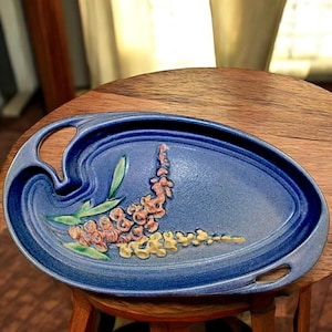May include: A blue ceramic serving tray with a floral design in shades of peach and yellow. The tray has a handle on one side and is oval-shaped. The tray is sitting on a wooden surface.