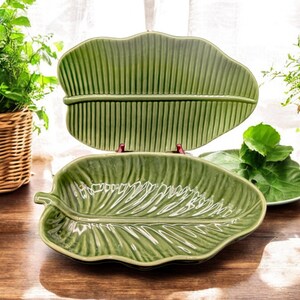 May include: Two green ceramic serving dishes shaped like banana leaves. The dishes are on a wooden table.