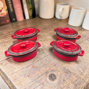 May include: Four small, red oval-shaped cocottes with lids and side handles. The lids have a silver rim and a red knob. The cocottes are arranged on a wooden surface, suggesting a kitchen or dining setting.