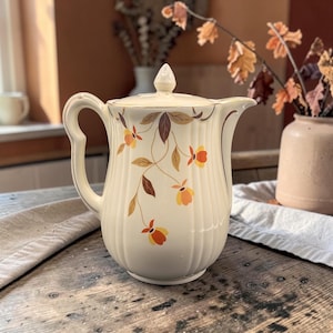 May include: A cream-colored ceramic coffee pot featuring a vertical ribbed texture and a delicate floral pattern in shades of orange and brown. The pot includes a matching lid with a pointed finial, a curved handle, and a gold-trimmed spout.