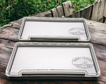 May include: Two clear plastic fly fishing boxes with white foam inserts. The boxes have a black logo that says "Harry Goodes Outfitters".