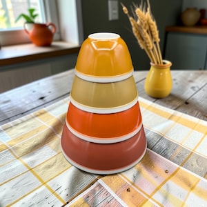 May include: A stack of four nesting bowls in graduated sizes, featuring a smooth finish and a warm color palette ranging from golden yellow at the top to a deep terracotta red at the base, with each bowl having a distinct white rim detail.