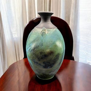 May include: A handcrafted ceramic vase with a wide, rounded body and a narrow neck. The vase features a blend of green and blue hues with dark accents. It has two small, curved handles near the top and stands approximately 12 inches tall.