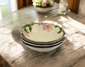 May include: A stack of four white ceramic bowls with a floral design. Each bowl features a pink flower with a yellow center and green leaves around the rim. The bowls are stacked on a wooden table.