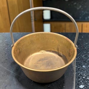 May include: A small, round, metallic pot with a brass-toned finish and a weathered, aged appearance. It features a simple, arched silver-colored metal handle attached to the sides, with visible signs of wear and oxidation on the interior surface.