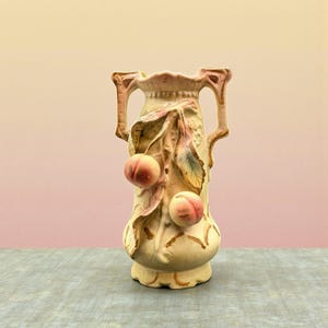 May include: A cream-colored ceramic vase with two handles and peach and leaf detailing. The vase features two peaches and several leaves in shades of green, pink, and gold. The vase is on a gray surface with a pink and yellow background.