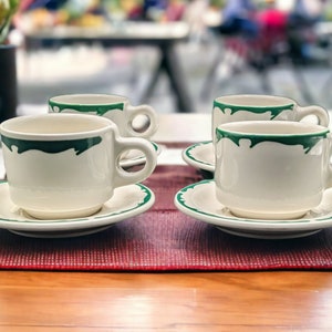 May include: Four white ceramic coffee cups with green trim and matching saucers on a red placemat. The cups are arranged in a square pattern.