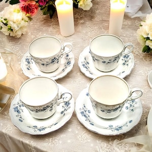 May include: Four white teacups with blue floral patterns and matching saucers. The cups and saucers are arranged on a white tablecloth with two white candles in the background.