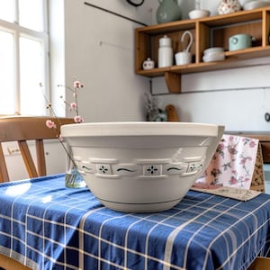 May include: A large, off-white ceramic mixing bowl featuring a raised, embossed basket-weave pattern around the exterior. The design is accented with small, dark green floral and wave-like motifs. The bowl has a wide, smooth rim and a rounded base.