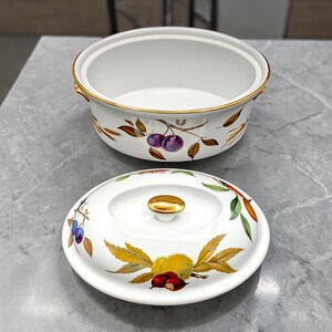 May include: White ceramic serving dish with a lid, featuring a gold rim and handle. The dish is decorated with a botanical design of plums, leaves, and chestnuts. The lid is placed next to the dish.
