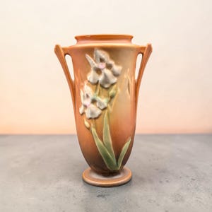 May include: A ceramic vase with a warm orange and brown gradient finish, featuring two side handles and a raised relief design of white iris flowers with green stems and leaves. The vessel has a circular base and a wide, open top rim.