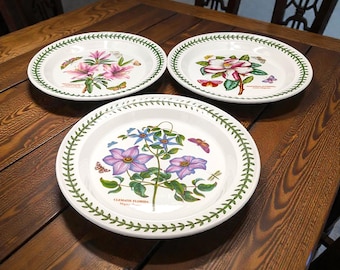 May include: Three white ceramic plates with a green leaf border pattern. Each plate features a unique botanical illustration of flowers and butterflies. The bottom plate shows Clematis Florida, while the top plates display Rhododendron and Magnolia.