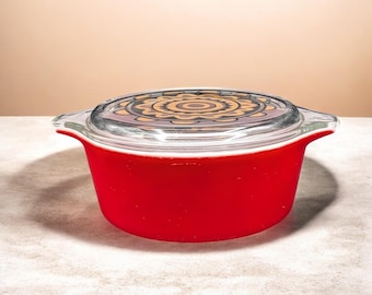 May include: A red Pyrex casserole dish with a clear glass lid. The lid features a brown and yellow floral design. The dish has two handles and is sitting on a light-colored surface. A vintage kitchenware item.