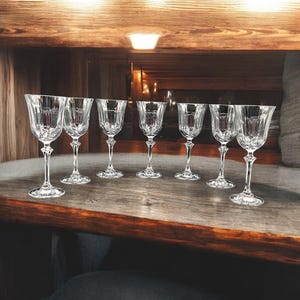May include: A set of seven clear crystal wine glasses with intricate cut patterns. The glasses have long stems and sit on a wooden surface, reflecting light. The glasses are arranged in a row, suggesting a formal setting.