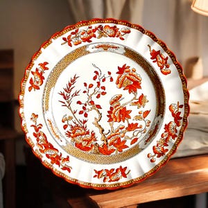 May include: A white ceramic plate with a scalloped edge, featuring a floral design in shades of red, orange, and brown. The plate has a decorative border and a central floral motif, suitable for serving food.