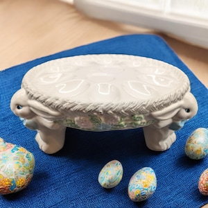 May include: A white ceramic Easter egg holder shaped like a bunny with a nest on top. The holder has six indentations for eggs and is decorated with floral details. The holder is sitting on a blue surface with several decorated Easter eggs.