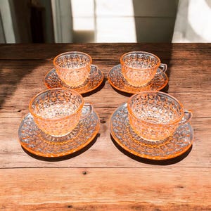 May include: Four vintage, clear glass teacups and saucers with an orange tint. The cups and saucers have a diamond pattern. The set is arranged on a wooden surface, with natural light casting shadows.