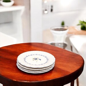 May include: A stack of four white ceramic plates with a blue floral design and a dotted border. The plates are stacked on a round, dark wooden table. The plates are in focus, while the background is blurred.