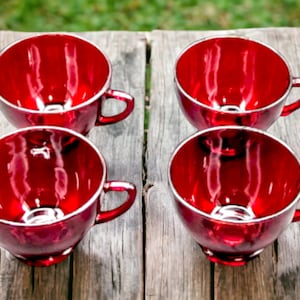 May include: Four red teacups with a shiny finish sit on a wooden surface. The cups are arranged in two rows of two.