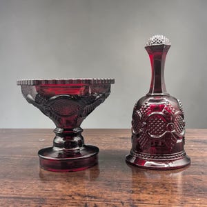 May include: A deep red glass pedestal bowl and a matching hand bell. Both items feature a pressed glass pattern with diamond-shaped textures and arched motifs. The bowl has a wide, scalloped rim and a sturdy base, while the bell has a tall handle.
