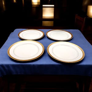 May include: Four white oval plates with gold rims and a thin gold line border are arranged on a blue tablecloth. The plates have a simple, elegant design, suitable for formal dining.