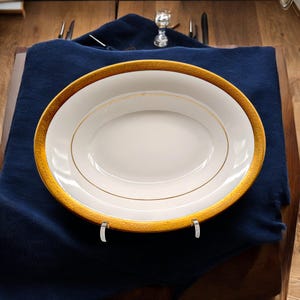 May include: An oval, white serving platter with a gold patterned rim and a gold inner border. The platter is displayed on a dark blue cloth, with silverware and a small glass visible in the background. The platter is supported by a clear stand.