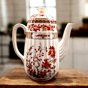 May include: A white ceramic coffee pot with a lid, handle, and spout. The pot is decorated with red and gold floral patterns and a decorative band. The pot is sitting on a wooden surface.