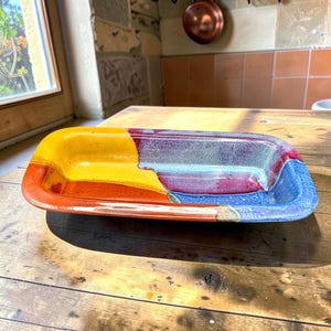 May include: A rectangular ceramic dish featuring a vibrant, multi-colored glaze. The surface displays distinct sections of bright yellow, deep orange, rich purple, and cool blue, blending together with a glossy, smooth finish and rounded edges.