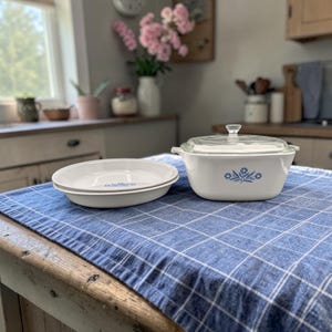 May include: White ceramic casserole dish with a glass lid and two white pie plates. Each piece features a blue floral design. The dishes are set on a blue and white checkered tablecloth in a kitchen setting.