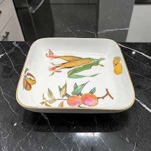 May include: A square, white ceramic serving dish with a gold rim. The dish is decorated with illustrations of corn, peaches, and other fruits. The artwork is in a botanical style, with vibrant colors and detailed depictions of the fruits and leaves.