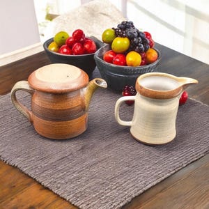 May include: A ceramic teapot with a lid and a small creamer pitcher sit on a gray placemat. The teapot is brown with horizontal stripes. The creamer is off-white with a brown rim. Bowls of fruit are in the background.