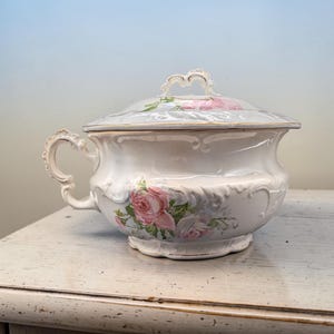 May include: A white ceramic chamber pot with a matching lid, featuring a decorative handle and embossed scrollwork details. The surface is adorned with a floral pattern of pink and white roses with green leaves and gold trim along the edges.