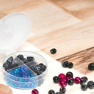May include: A clear plastic container with four compartments filled with colorful beads. The container has a lid and is sitting on a wooden surface. There are also loose beads scattered around the container.