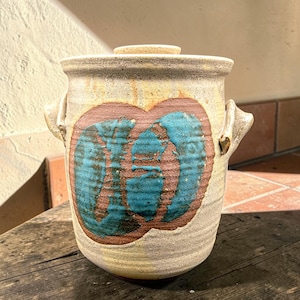 May include: A cylindrical ceramic jar with a lid and two side handles. The exterior features a speckled beige and tan glaze with a prominent circular design in the center, showcasing a textured brown ring filled with vibrant turquoise blue glaze.