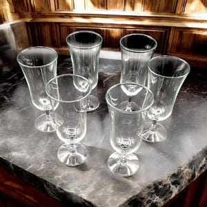May include: Six clear glass goblets with a stemmed base are arranged on a dark gray countertop. The glasses have a tulip-shaped bowl and a flared rim. The background features dark wood cabinetry.