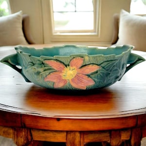 May include: A teal ceramic bowl with a floral design. The bowl features a large, stylized flower in shades of pink and yellow at the center. The bowl has handles and is placed on a wooden table. The background includes a window and pillows.