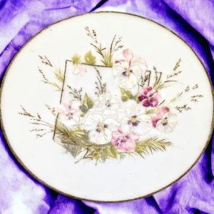 May include: A white ceramic plate with a gold rim features a hand-painted floral design. The design includes white, pink, and purple pansies with green leaves and stems.