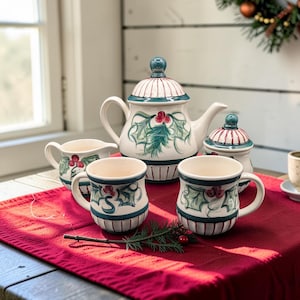 May include: A ceramic tea set featuring a teapot, a lidded sugar bowl, a creamer, and two mugs. Each piece is cream-colored with hand-painted green holly leaves, red berries, and dark green bands. The teapot and sugar bowl have matching lids.
