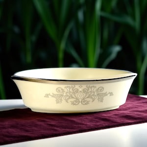 May include: A white oval serving bowl with a silver rim and a delicate floral design. The bowl is sitting on a burgundy fabric surface.