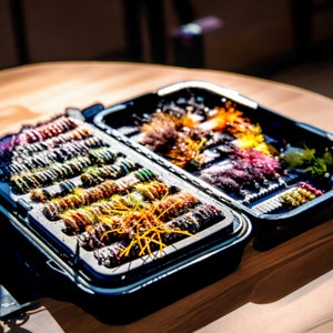 May include: A black fly fishing tackle box with two compartments filled with colorful fly fishing lures.