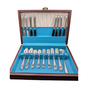 May include: A wooden storage case with a hinged lid containing a set of silver-toned flatware. The interior features a bright blue fabric lining with individual slots for four knives, four spoons, and four forks, arranged in a structured display.