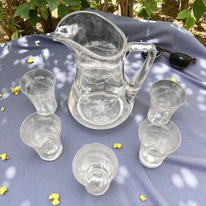May include: A clear glass pitcher with a handle and six matching glasses. The pitcher and glasses have etched floral designs. The set is arranged on a blue surface, with sunglasses in the background.