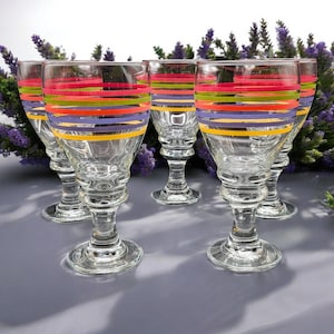 May include: Set of six clear wine glasses with colorful horizontal stripes in red, green, orange, purple, and yellow. The glasses have a classic stemmed design, perfect for serving wine or other beverages.