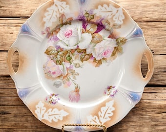 May include: A white porcelain serving platter with two handles, featuring a floral design of pink and white roses with green leaves. The platter has a scalloped edge and a light brown rim.