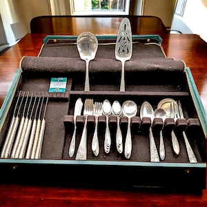 May include: A vintage silver flatware set displayed in a green and brown velvet-lined case. The set includes knives, forks, spoons, and serving utensils. The silverware has ornate floral detailing and is arranged neatly within the case.